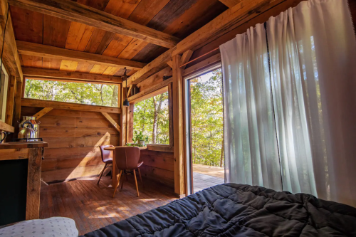 Cozy wooden cabin interior with a bed, dining area, and large windows overlooking a forest. Natural light floods the space.