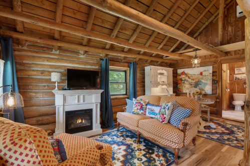Cozy log cabin interior with a fireplace, patterned sofas, and rustic decor, featuring wooden beams and a colorful rug.