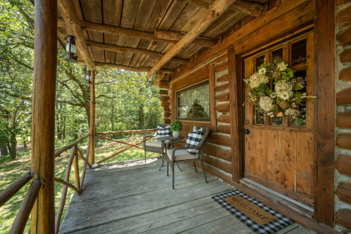 A rustic wooden porch with two chairs, a welcome mat, and a floral wreath on the door, surrounded by trees.