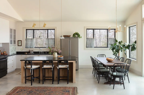 Modern kitchen with an island, dining table, large windows, and plants, featuring a mix of black and wooden furniture.