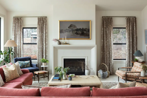 Cozy living room with a fireplace, pink sofas, patterned chairs, and large windows, adorned with plants and artwork.