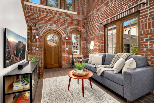 Cozy living room with brick walls, a gray sofa, wooden door, and a round table on a patterned rug.