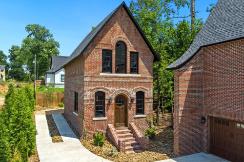 A charming brick house with arched windows and a wooden door, surrounded by greenery and a clear blue sky.