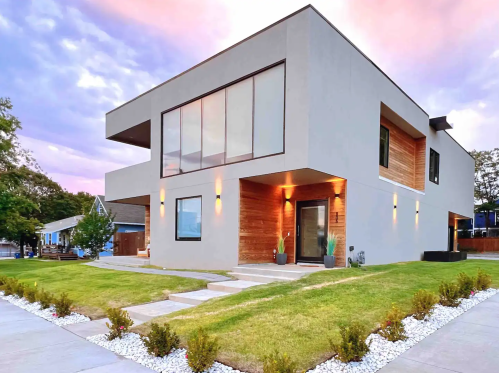 Modern two-story house with large windows, wooden accents, and landscaped yard under a colorful sky.