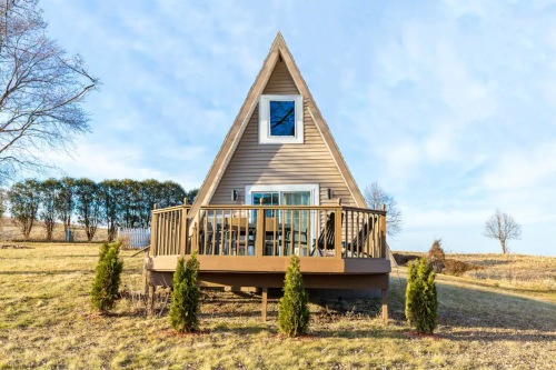 A cozy A-frame cabin with a deck, surrounded by grass and small trees, under a clear blue sky.