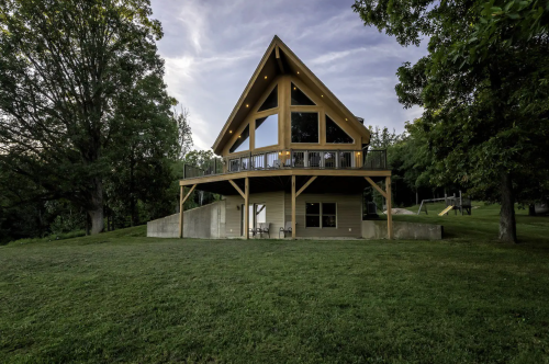 A modern A-frame house with large windows, surrounded by trees and a grassy area, under a cloudy sky.