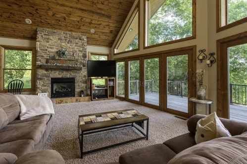 Cozy living room with stone fireplace, large windows, and a view of the outdoors, featuring comfortable seating and a coffee table.