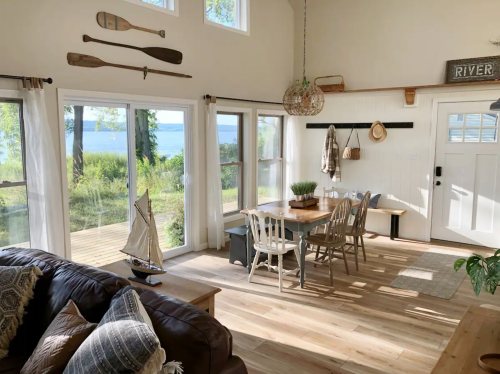 Bright, airy living space with a dining table, chairs, and views of the river through large windows. Decor includes paddles.
