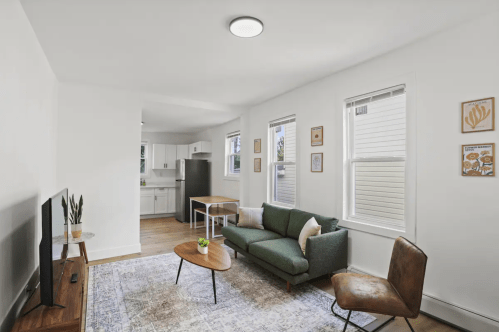 Cozy living room with a green sofa, wooden coffee table, and kitchen in the background, featuring bright natural light.