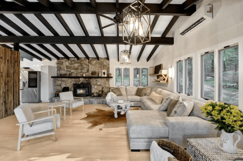 Cozy living room with a stone fireplace, large sectional sofa, and wooden beams, surrounded by natural light.