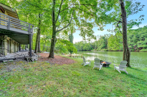 A serene lakeside scene with a fire pit, Adirondack chairs, and lush green trees beside a calm water body.