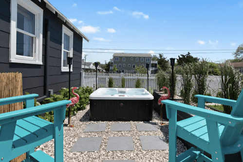 A backyard with a hot tub, turquoise chairs, flamingo decorations, and a white picket fence under a clear blue sky.