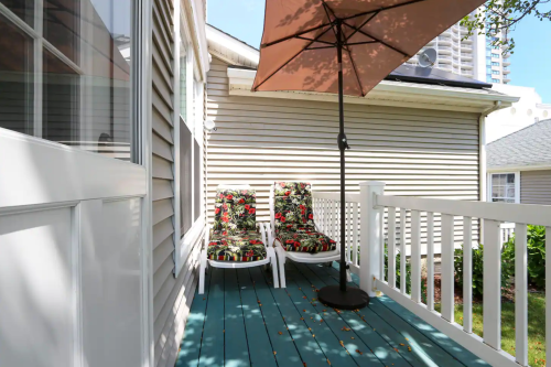 Two colorful lounge chairs under an umbrella on a sunny deck, surrounded by light-colored siding and greenery.