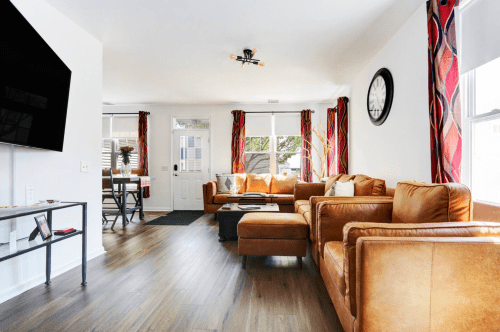 Bright living room with brown leather sofas, a coffee table, and large windows, featuring colorful curtains and a wall clock.