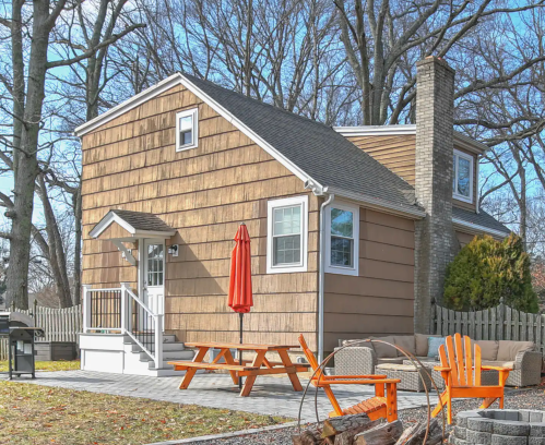A cozy brown-shingled house with a red umbrella on the patio, surrounded by trees and outdoor seating.