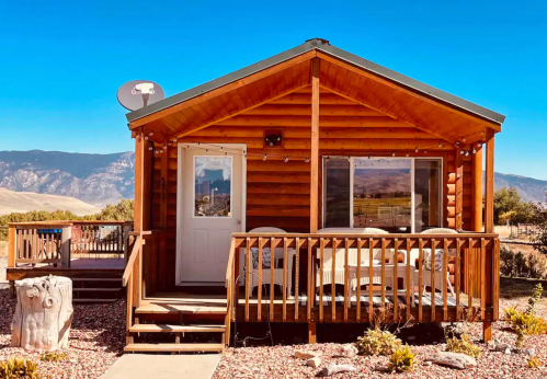 A cozy wooden cabin with a porch, surrounded by mountains and clear blue skies.