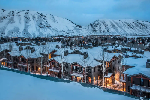 Snow-covered mountains loom over a cozy, illuminated village nestled among trees in a winter landscape.