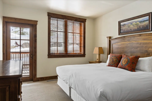 Cozy bedroom with a wooden bed, decorative pillows, a lamp, and a door leading to a balcony with snowy views.