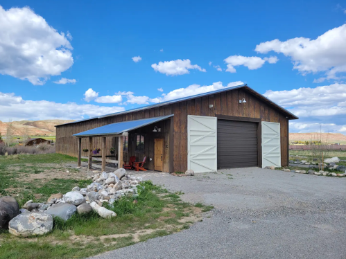 A rustic wooden barn with a metal roof, surrounded by rocks and grass, under a blue sky with fluffy clouds.