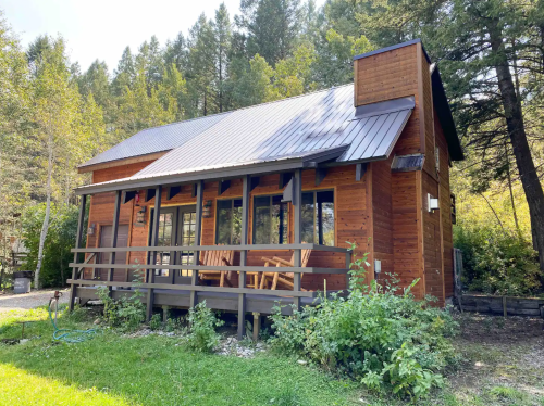 A cozy wooden cabin with a metal roof, surrounded by trees and greenery, featuring a front porch with chairs.
