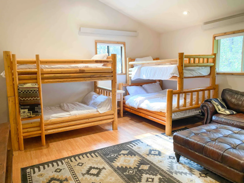 Cozy room with two sets of wooden bunk beds, a leather couch, and a patterned rug. Natural light from windows.