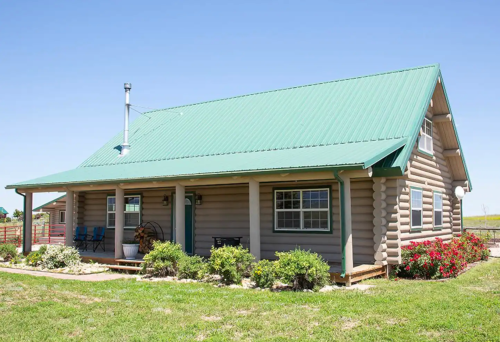 A rustic log cabin with a green metal roof, surrounded by greenery and flowers, under a clear blue sky.