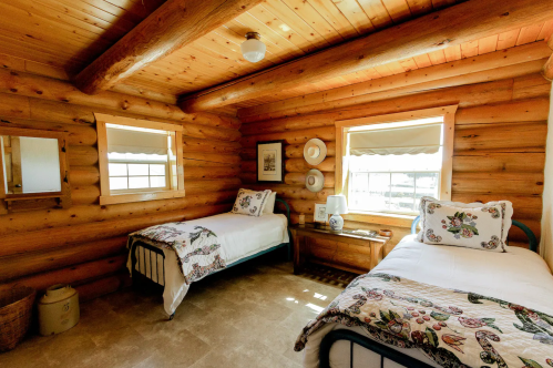 Cozy log cabin bedroom with two beds, wooden walls, and natural light from windows. Decorated with floral quilts.
