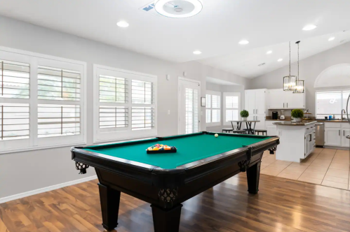 A bright room featuring a green pool table, large windows, and a modern kitchen in the background.
