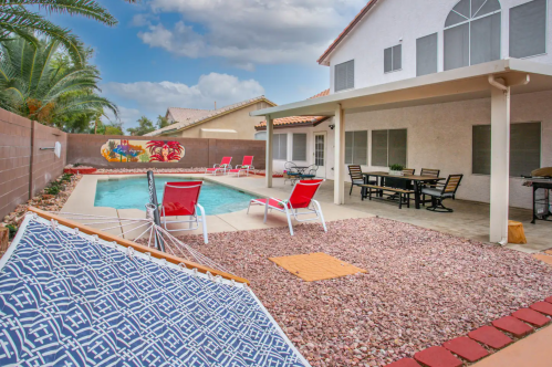 Backyard with a pool, lounge chairs, a hammock, and a dining area under a covered patio, surrounded by palm trees.