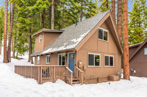 A cozy brown cabin surrounded by snow and tall trees, featuring a deck and large windows.