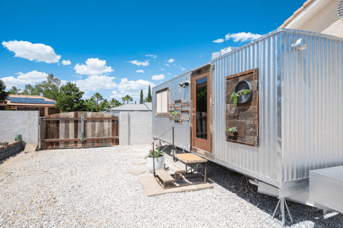 A modern tiny home with a metal exterior, steps leading up, and a gravel yard under a blue sky with clouds.