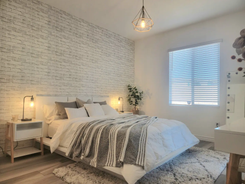 Cozy bedroom with a white brick wall, a bed with gray blankets, and a window with blinds, featuring modern decor.