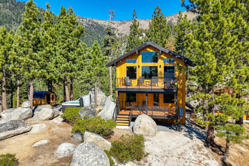 A modern wooden cabin surrounded by tall trees and large boulders, set against a mountainous backdrop.