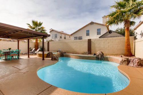A backyard pool surrounded by palm trees, a patio area with seating, and houses in the background under a cloudy sky.