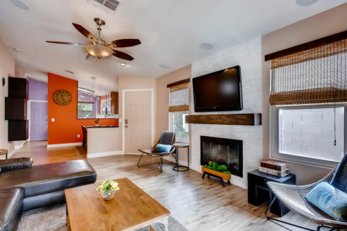 Cozy living room with a ceiling fan, TV above a fireplace, and modern furniture, featuring warm colors and natural light.