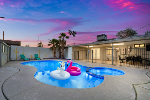 A pool with inflatable swan and flamingo floats, surrounded by palm trees and a colorful sunset sky.
