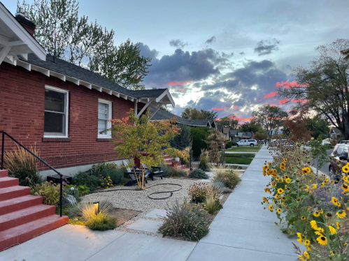 A peaceful neighborhood scene with brick houses, colorful gardens, and a sunset sky filled with clouds.
