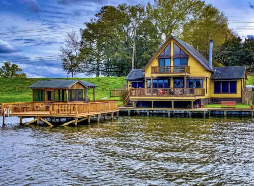 A large yellow house with a deck by the water, featuring a wooden dock and a small gazebo nearby, under a cloudy sky.