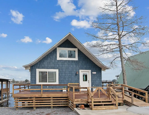 A blue, A-frame house with a wooden deck, surrounded by a clear sky and a few clouds.