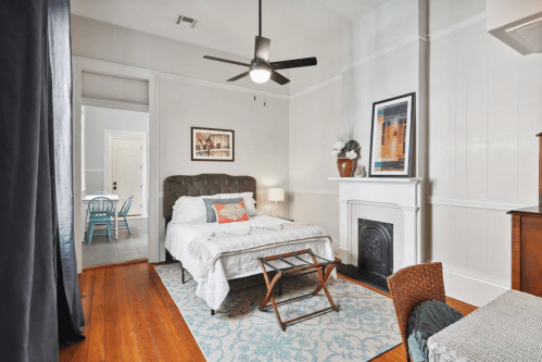 Cozy bedroom with a bed, fan, and decorative fireplace, featuring a small table and a view of a dining area.