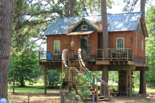 A charming wooden treehouse with a metal roof, featuring a spiral staircase and surrounded by trees.