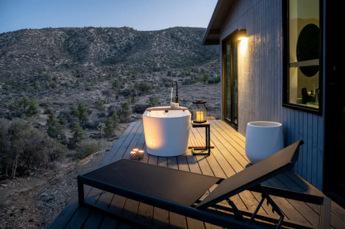 A modern outdoor deck with a freestanding bathtub, lounge chair, and scenic mountain view at dusk.