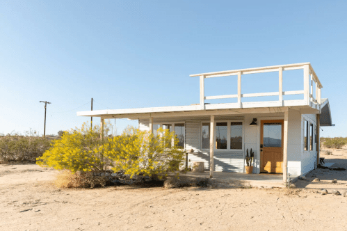 A modern, single-story house with a rooftop deck, surrounded by desert vegetation and clear blue skies.