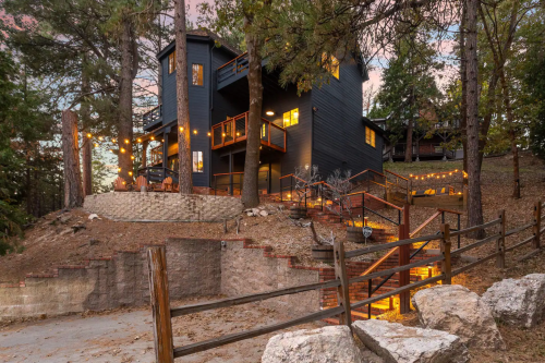 A modern black house nestled among trees, featuring outdoor lighting and a staircase leading to a cozy patio area.