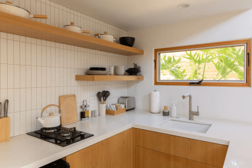 Modern kitchen with wooden shelves, white countertops, and a window overlooking greenery. Minimalist decor and utensils.