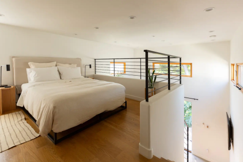 A modern bedroom with a white bed, wooden floors, and a railing overlooking a bright, open space.