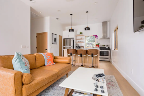 Modern living room with a brown leather sofa, a white coffee table, and a kitchen with bar stools in the background.