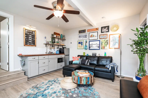Cozy living room with a black sofa, wooden coffee table, wall art, and a small kitchenette in a bright, modern space.