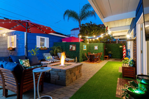 Cozy outdoor patio with a fire pit, seating, and string lights, surrounded by greenery and colorful umbrellas at dusk.