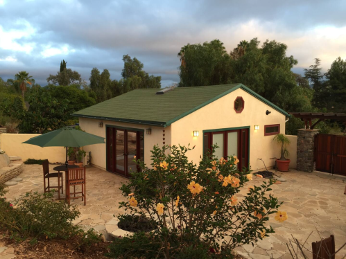 A cozy house with a green roof, surrounded by trees and flowers, featuring a patio with outdoor furniture.